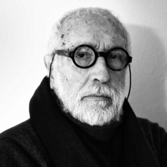 Andrea Branzi portrait, grayscale photograph, elderly man with white beard wearing black turtleneck and glasses, contemplative expression.