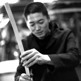 Arihiro Miyake portrait holding rectangular wooden staff, dark turtleneck, blazer, blurred interior background, soft diffused lighting.