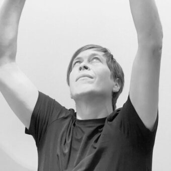 Black and white portrait of Bernhard Osann, a man with short brown hair, gazing upwards while raising his arms, against a light gray background.