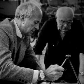 Black and white photograph of two men examining a small metallic object on a table, wearing tweed and dark shirts.
