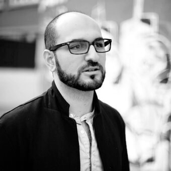 Charles Kalpakian portrait, black jacket, dark glasses, light shirt, contemplative expression, blurred abstract background, studio lighting.