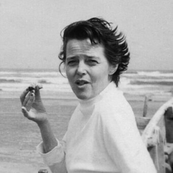 Charlotte Perriand portrait, black and white photograph, beach scene, white shirt, short hair, holding rectangular object, ocean background.