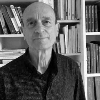 Grayscale portrait of a man between bookshelves, displaying book titles including “Hard Times” and Italian literature, wearing a dark textured shirt.