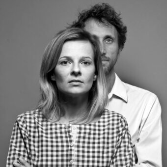 Gray scale portrait of a couple, woman in checkered shirt, man in white shirt, both with direct gazes, studio lighting, “euga design” visible.