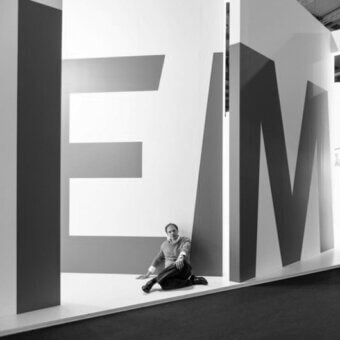 Black and white graphic installation of “IEM” letters, white panels, gray forms, man seated, studio setting, geometric composition, minimalist aesthetic.