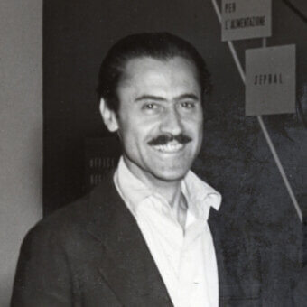Black and white portrait of Franco Albini, a man in a suit smiling at the camera, with Italian signage visible in the blurred background.