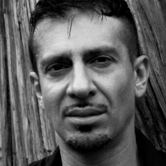 Black and white portrait of Gabi Peretto with short dark hair, goatee, and neutral expression against a weathered wood plank background.