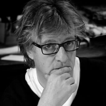 Javier Mariscal portrait, black and white, designer with glasses, hand to chin, white shirt, black sweater, blurred studio background.