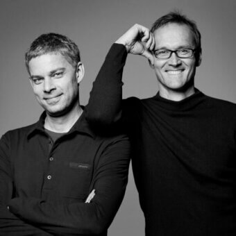 Black and white portrait of two men, designers Jehs Laub, wearing dark clothing, posed in a gray studio background with relaxed expressions.