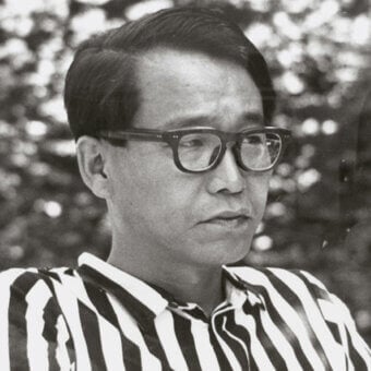 Black and white portrait of Kazuhide Takahama, a man with short dark hair, wearing striped shirt and glasses, contemplative expression.