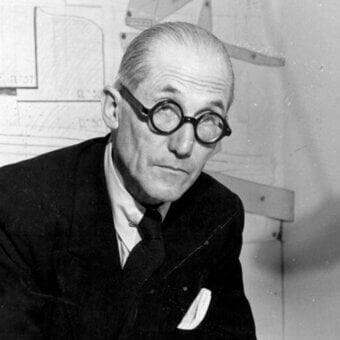 Le Corbusier portrait, black and white, wearing a suit and glasses, with blurred architectural blueprints and technical drawings in background.