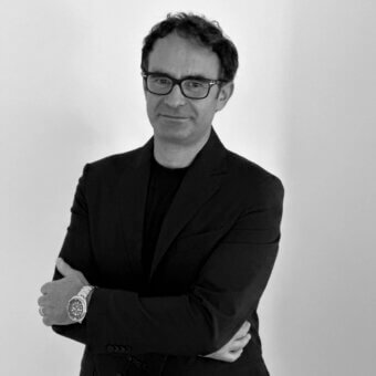 Man in black suit and glasses, silver wristwatch, crossed arms, neutral expression, plain light gray background, portrait.
