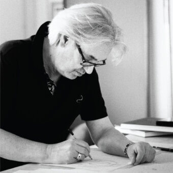 Marco Innocenti drawing technical sketch, white hair, glasses, black polo, workspace, soft lighting, silver ring, metal watch.