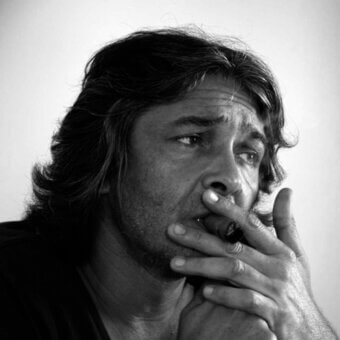 Black and white portrait of Rudy Ricciotti with beard smoking a cigar, soft lighting, contemplative expression, gray background.
