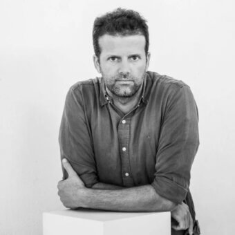 Man in olive shirt, black and white portrait, arms crossed on white cube, direct gaze, minimalist composition, diffused lighting, textured hair.