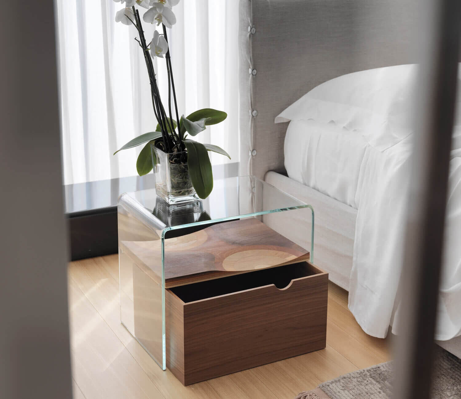 Cassoni Horm Bifronte bedside table, dark wood base, rectangular shape with glass top displaying white orchids in a vase, modern bedroom setting.