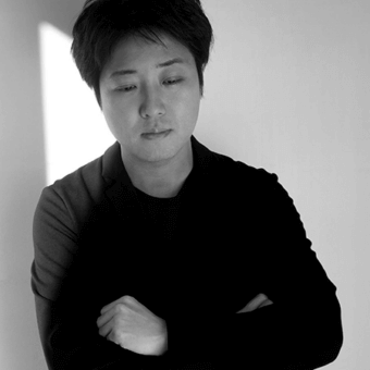 Black and white portrait of a person with short dark hair, wearing a dark crewneck sweater, arms crossed, against a white wall with vertical shadow.