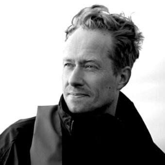 Black and white portrait of Esa Vesmanen, facing right, wearing a dark high-collared jacket, with textured hair and a contemplative expression.