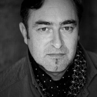 Black and white portrait of Jean-François Gomrée, dark jacket, geometric scarf, direct gaze, soft lighting, detailed facial features.