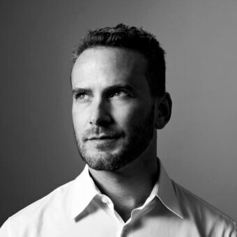 Black and white portrait of Joe Doucet, a man with short dark hair and beard, wearing a white button-down shirt, against a gradient gray background.