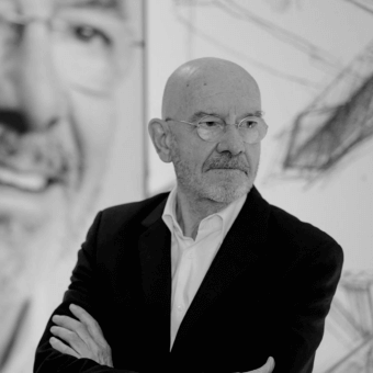 Mario Bellini portrait, black and white photograph, man in suit with crossed arms, artworks in blurred background, studio lighting.