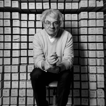 Architect Mario Botta portrait, archival boxes background, stacked grid pattern, numerical labels, neutral tones, diffused lighting.