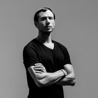 Black and white portrait of Sebastian Errazuriz, a man with dark hair, wearing a black v-neck shirt, against a gradient gray backdrop.