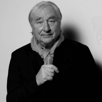 Black and white portrait of architect Steven Holl, wearing a dark jacket and light scarf, with direct eye contact and subtle expression.