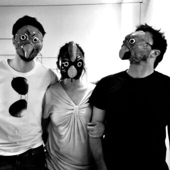 Black and white photograph of three people wearing textured bird masks with large eyes, set against a neutral wall.