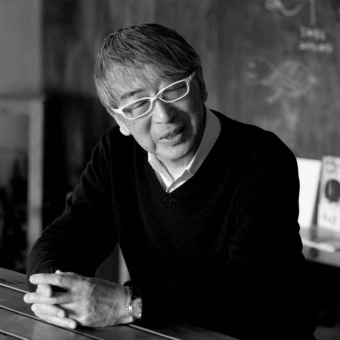Toyo Ito portrait, black and white photograph, man seated at wooden table wearing glasses and sweater, blurred background with text.