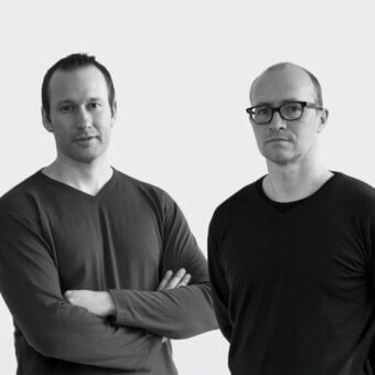 Black and white studio portrait of two men in black v-neck shirts, facing forward against a white backdrop, with neutral expressions.