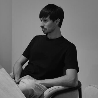 Black and white portrait of Federico Peri seated in an upholstered chair with a wooden frame, against a light gray wall.