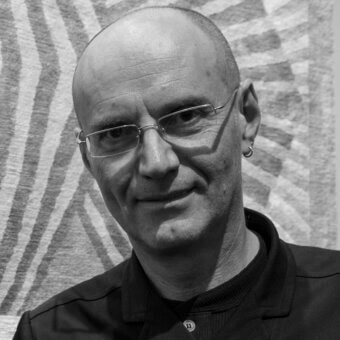 Bald man with glasses, black polo shirt, against gray zebra-patterned background, silver frame, hoop earring, soft lighting, portrait.