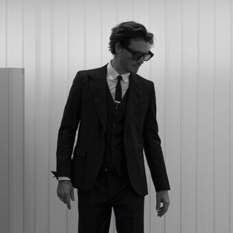 Gianmaria Conficconi portrait, man in black three-piece suit, rectangular glasses, against light wood panel backdrop, high contrast lighting.