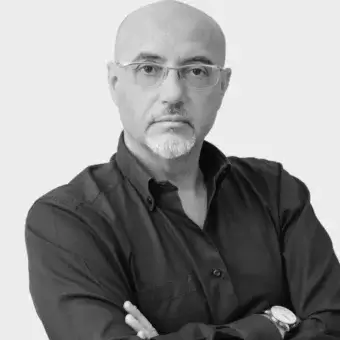 Bald man Giuseppe Bavuso in glasses, dark button-down shirt, gray goatee, crossed arms, wristwatch, light gray backdrop, portrait.
