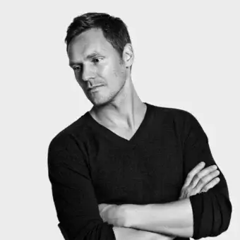 Black and white portrait of a man with short dark hair, wearing a dark v-neck sweater, arms crossed, against a light gray background.