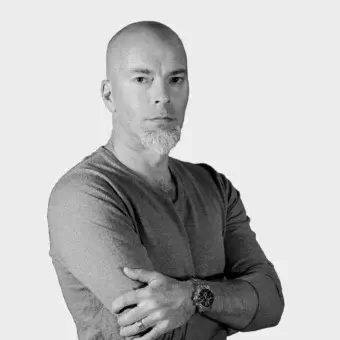 Black and white portrait of Karri Monni, a man with a shaved head and salt-and-pepper goatee, wearing a dark gray v-neck sweater and wristwatch.