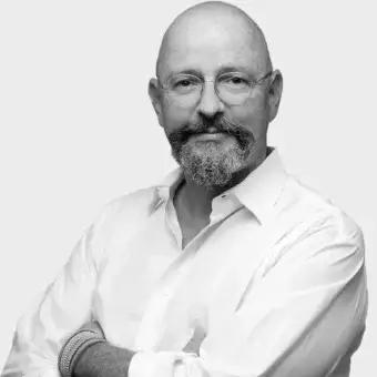 Black and white portrait of Klaus Nolting, a man with a gray beard and glasses, wearing a button-down shirt and woven bracelets.