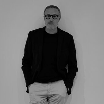 Marco Fumagalli portrait, man in black blazer and gray trousers, minimalist light gray background, short gray hair, wearing glasses.