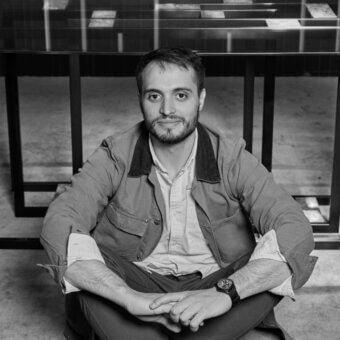 Matteo Bimbi portrait, man in tan jacket and dark trousers seated against geometric dark frame and translucent panel backdrop.