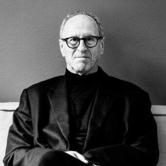 Matteo Thun portrait, gray hair and goatee, black turtleneck, dark blazer, seated on light beige upholstery against a gray wall.