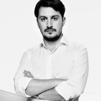 Man portrait, white shirt, dark hair, trimmed beard, silver wristwatch, neutral expression, high-key studio lighting, plain white background.