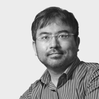Black and white portrait of Shin Azumi, a man with glasses, striped shirt, dark hair, and beard against a light gray background.