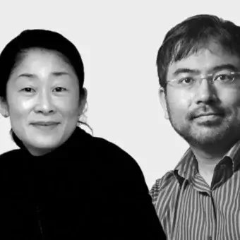 Black and white portrait of two people, Tomoko Azumi and a man with glasses, wearing casual clothing against a light gray backdrop.