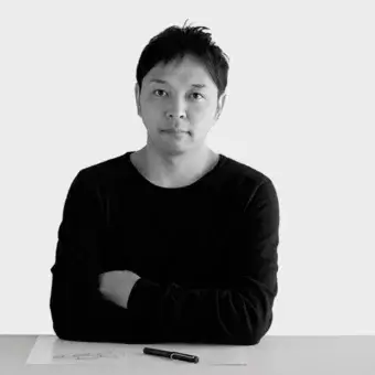 Black and white portrait of Tomoya Tabuchi, seated at a white table with a sketch, wearing a black long-sleeved shirt against a white wall.