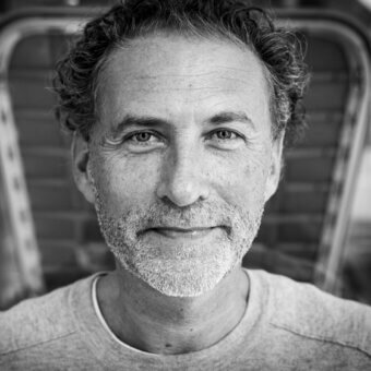 Werner Aisslinger portrait, black and white, curly hair, gray t-shirt, relaxed expression, soft lighting, close-up headshot, wooden backdrop.