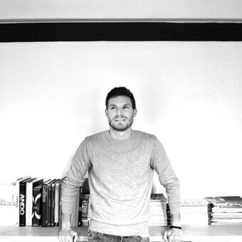 Black and white photograph of a man by a bookshelf filled with books and papers, wearing a gray sweater and jeans.