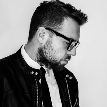 Man in black leather jacket, white shirt, and dark tie, wearing glasses, with short textured hair and beard, studio portrait.