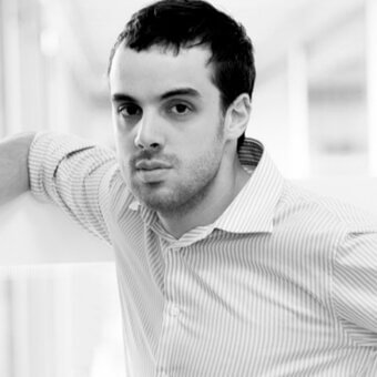 Black and white portrait of a man in a gray striped shirt leaning against a white architectural wall, neutral expression, blurred background.