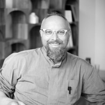 Black and white portrait of Giulio Iacchetti, a man with a beard wearing glasses and a button-down shirt, seated near bookshelves.
